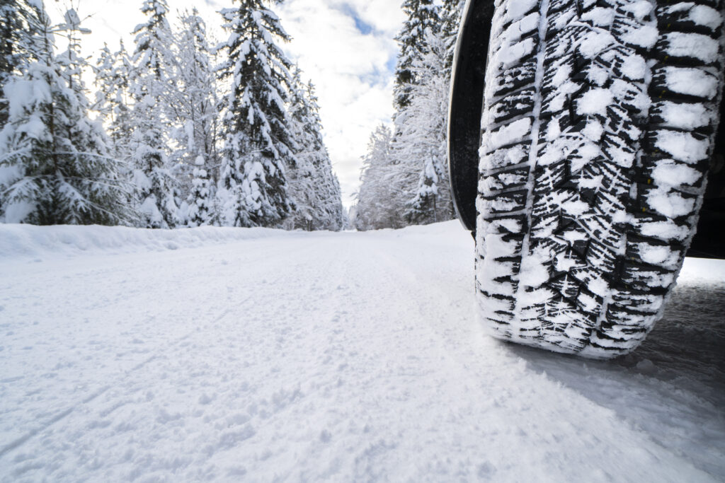 A winter tire on a motorhome, on a road that is full of snow. There is even more snow on the sides of the road and the pine trees are covered in snow as well. 