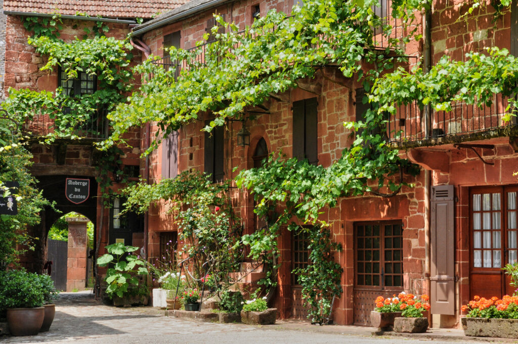 You see red-stoned houses and an archway, all covered with greenery. These red houses can be found in Collonges La Rouge in France